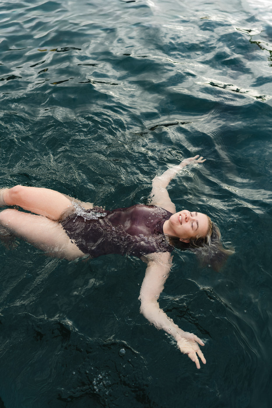 Dark one-piece swimsuit by Lilja the Label worn while floating on rippling water.