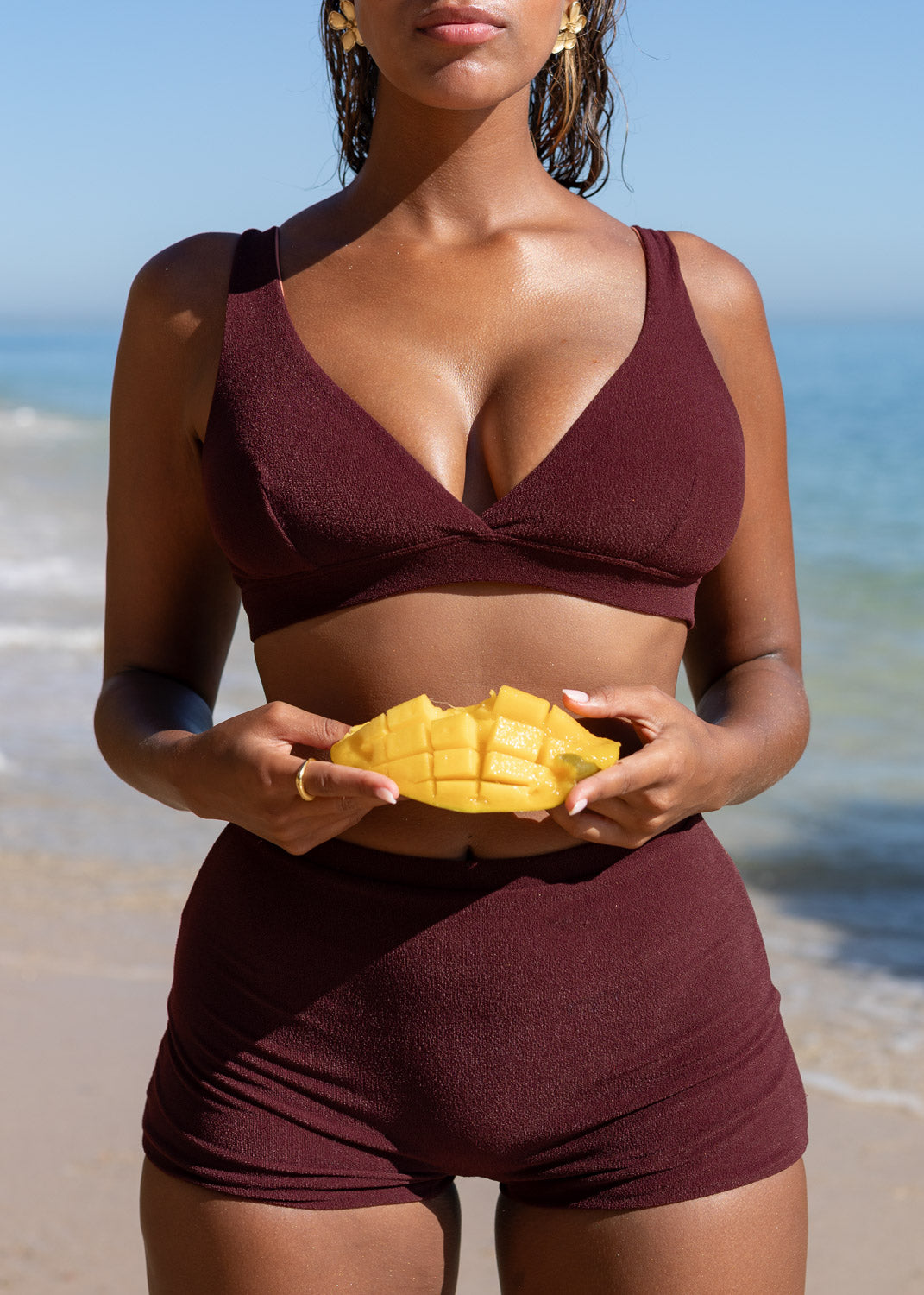 Wine red mini shorts by Lilja the Label, worn with a matching top, holding a cut mango at the beach.