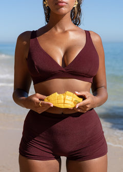 Wine red mini shorts by Lilja the Label, worn with a matching top, holding a cut mango at the beach.
