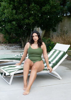 Olive ruched onepiece by Lilja the Label on a model sitting on a beach chair near green foliage.
