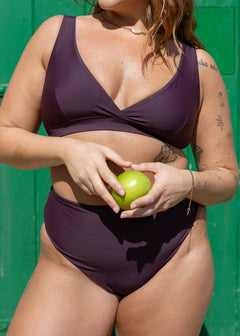 Plum high-leg bikini bottoms by Lilja the Label, held with a green apple, over a green background.