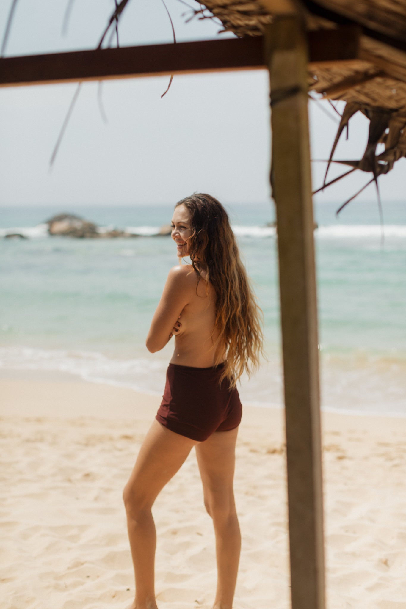 Sienna mini shorts in deep brown, high-waisted with smooth stretch fabric, shown on model from side view on sandy beach.
