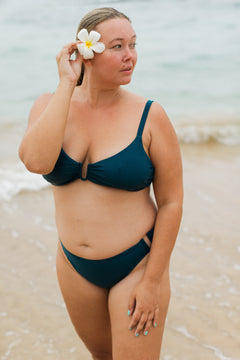Teal underwire bikini top with ruched center and thin straps, worn on beach with white flower in hair, side view